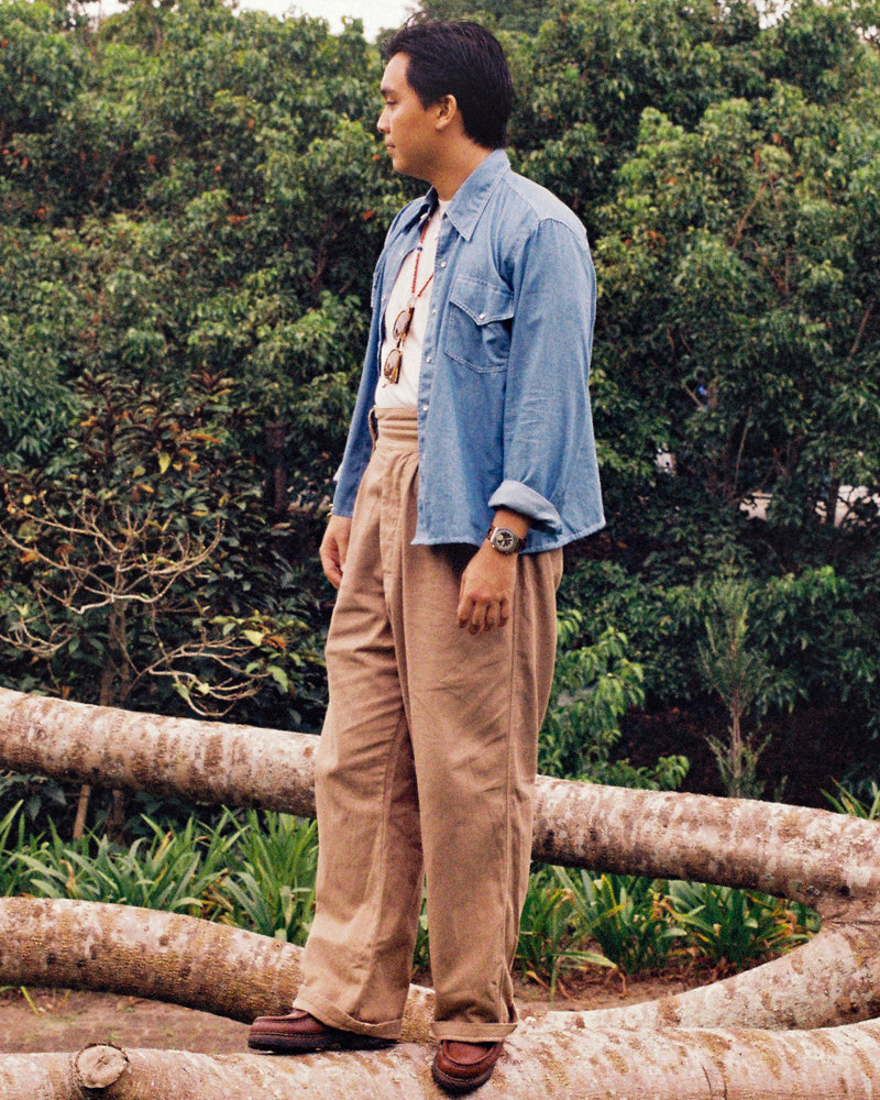 Person wearing a blue chambray shirt and beige pants standing outdoors with greenery in the background