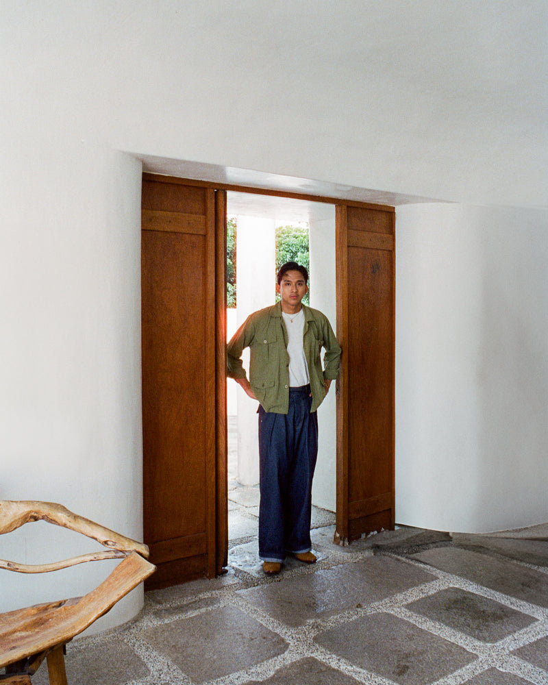 Person standing in a doorway with a wooden chair and white walls. Wearing a green safari shirt and denim gurkha pants. 
