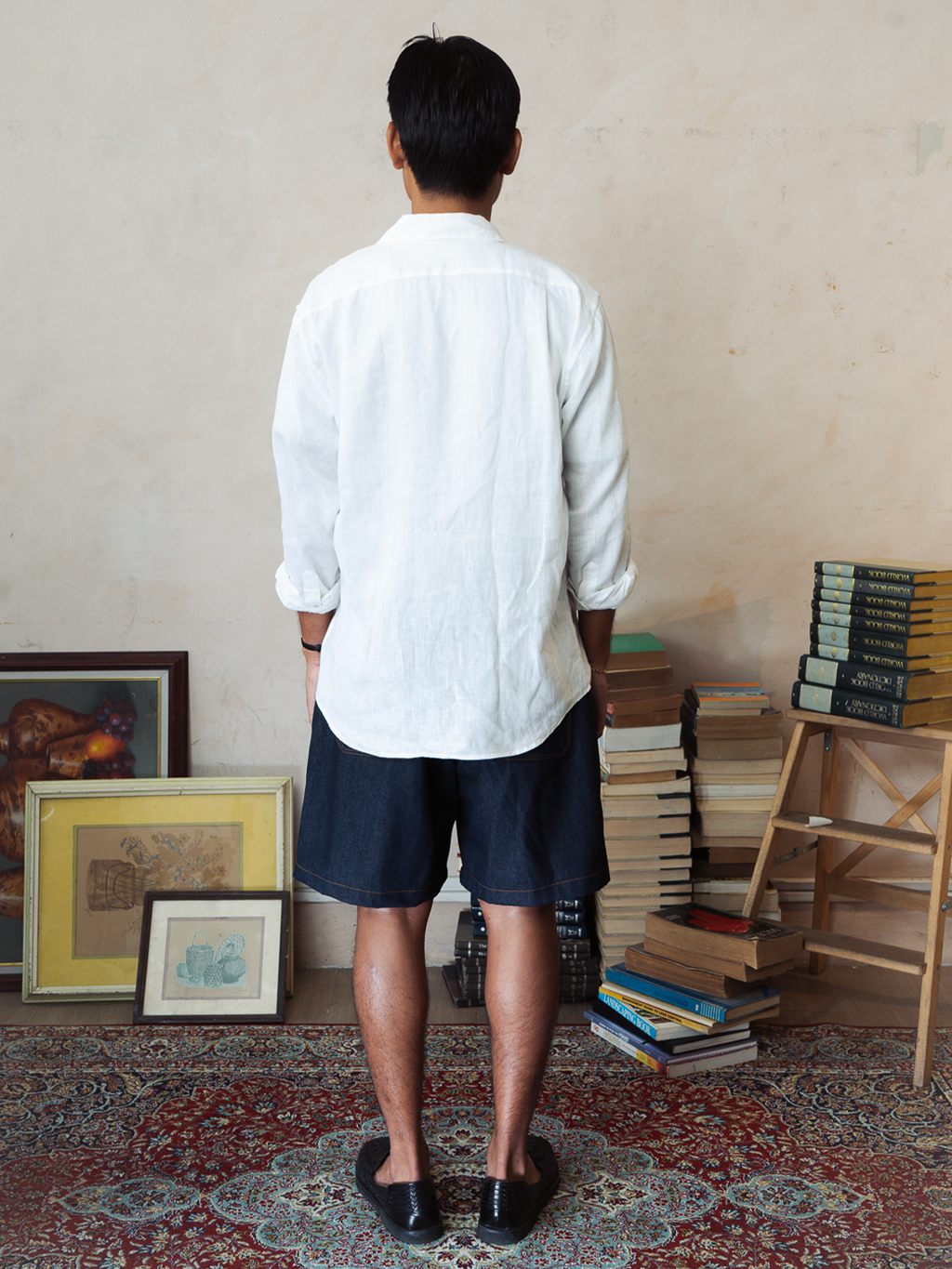 Man standing in a room with books and a wooden shelf. With Tous denim indigo shorts.