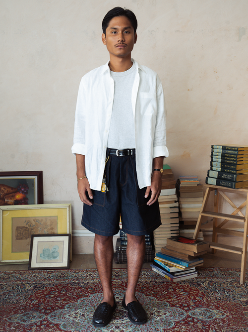 Man wearing a white shirt and tous dark denim shorts standing in a room with books and a rug.