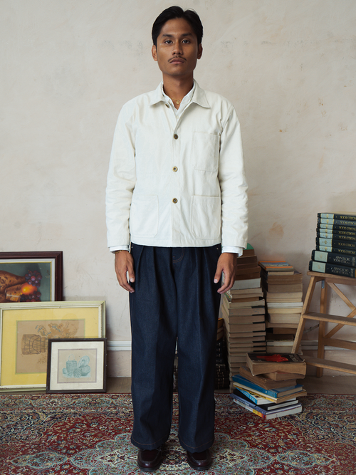 Man wearing a Cream canvas french chore jacket shirt and indigo gurkha pants standing in a room with books and pictures on shelves.