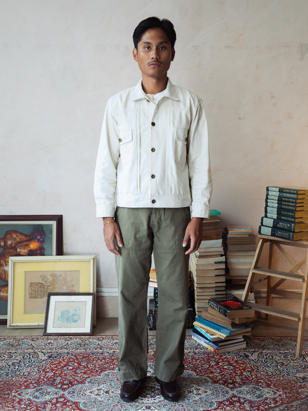 Man wearing a white tous type 2 trucker jacket and green pants standing in a room with books and artwork.
