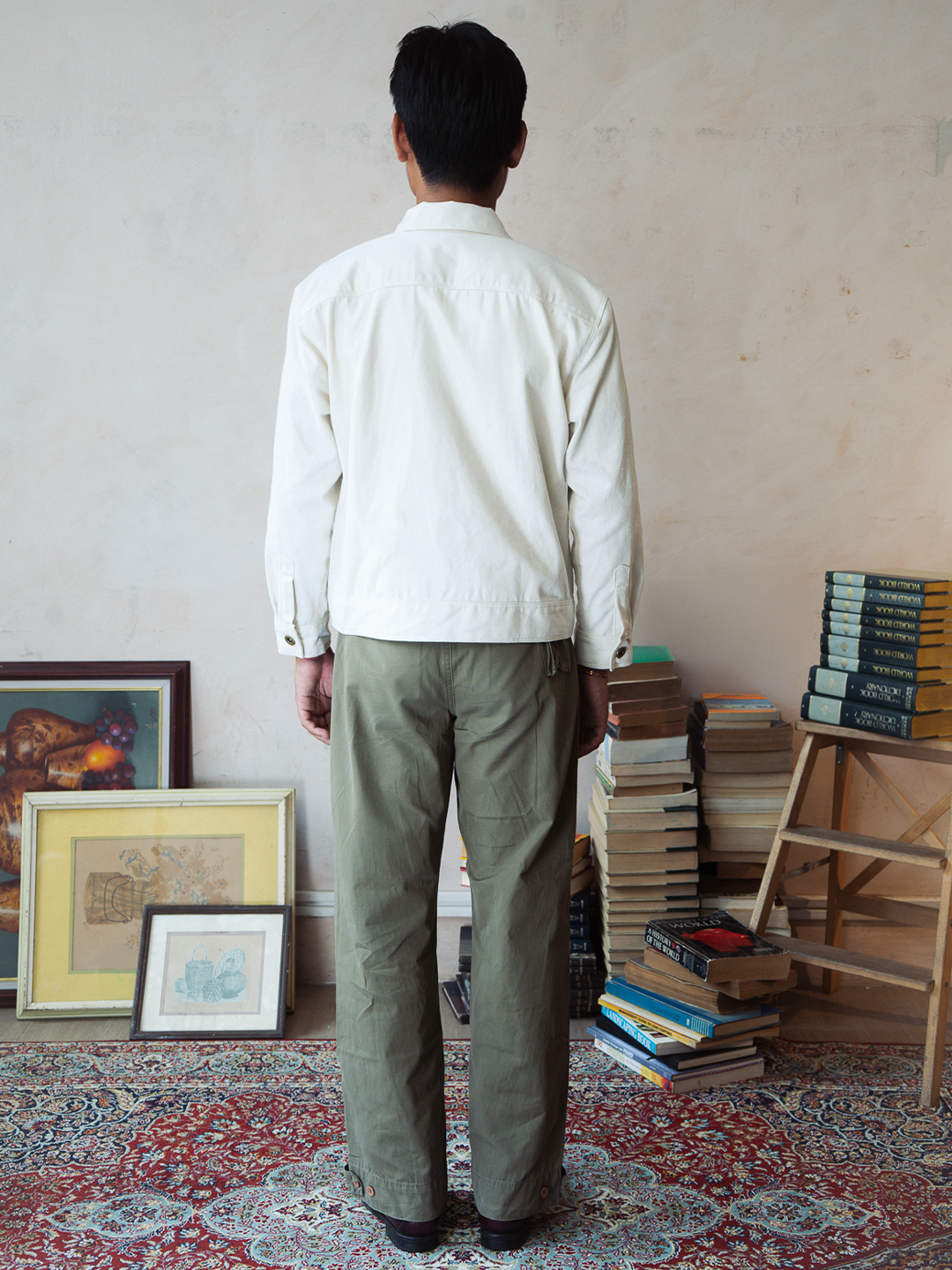 Person wearing a white type 2 trucker jacket and green pants standing in a room with books and framed pictures on a patterned rug.