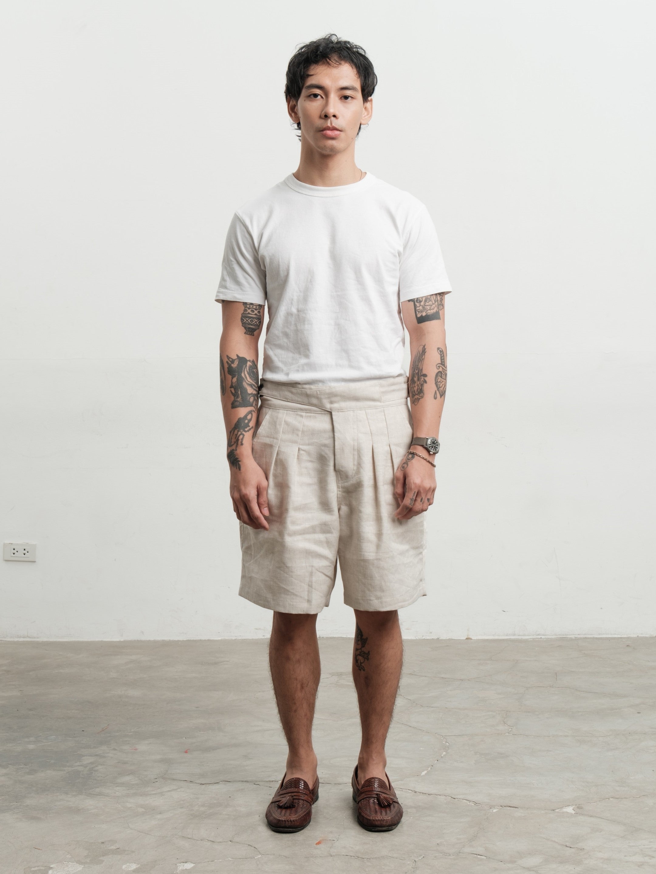 Person wearing a white t-shirt and cream pleated workwear gurkha shorts standing against a plain white wall.