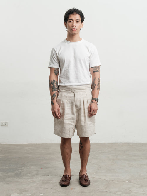 Person wearing a white t-shirt and cream pleated workwear gurkha shorts standing against a plain white wall.