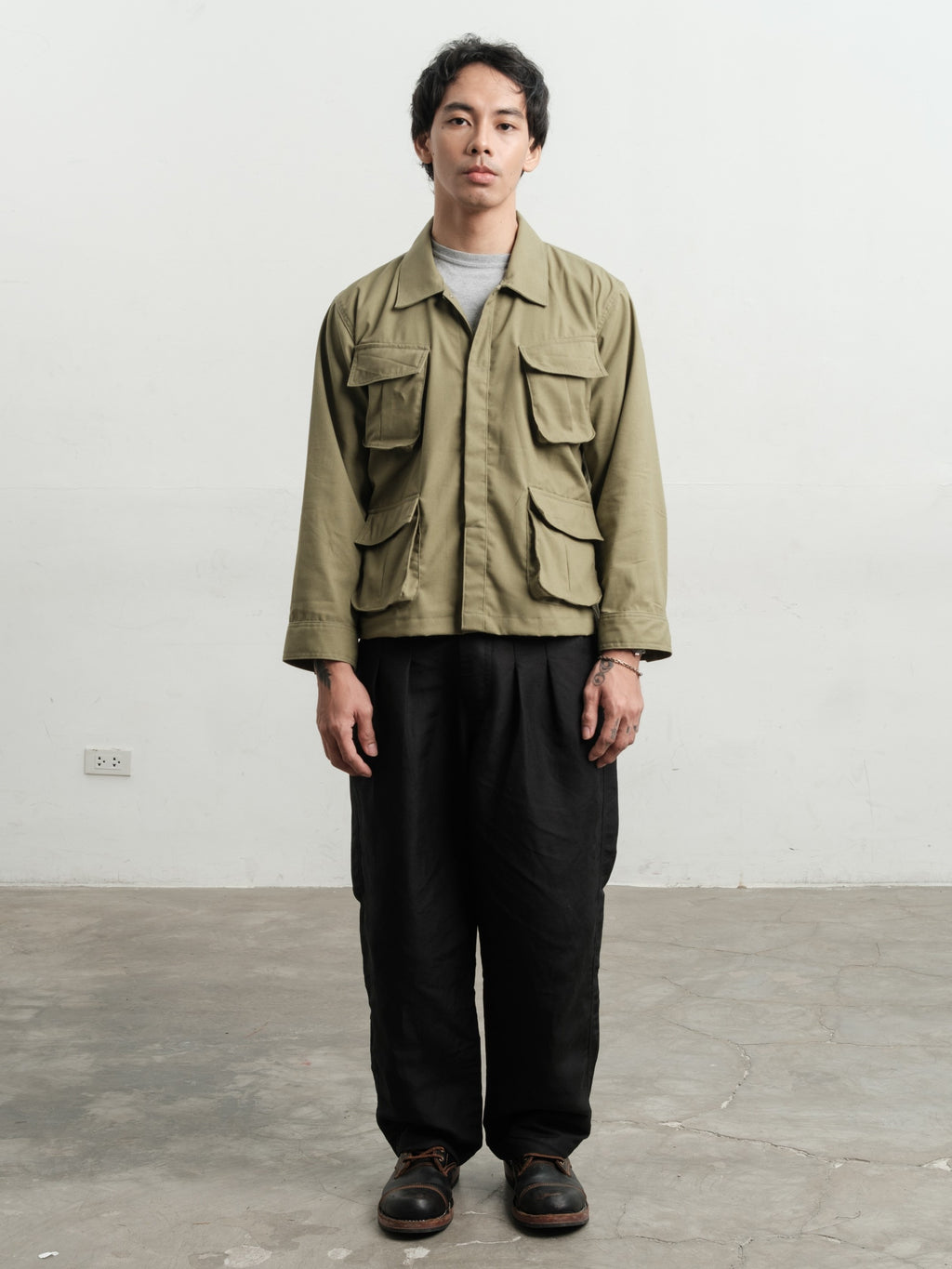 Man wearing a fatigue linen jungle jacket and linen black gurkha pants standing against a plain white wall.