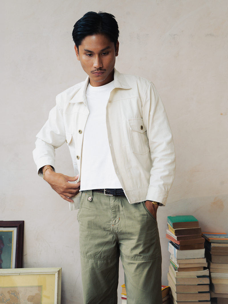 Man wearing a cream trucker jacket and green pants standing in front of a stack of books and framed pictures.