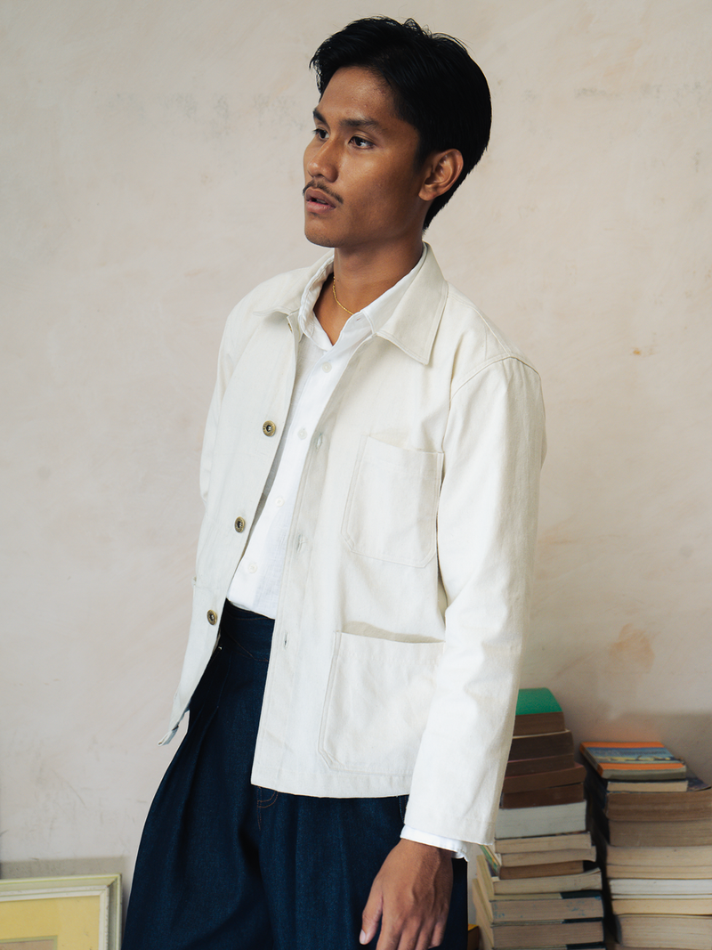 Person wearing a tous cream canvas jacket over a white shirt with dark pants, standing in front of stacked books.