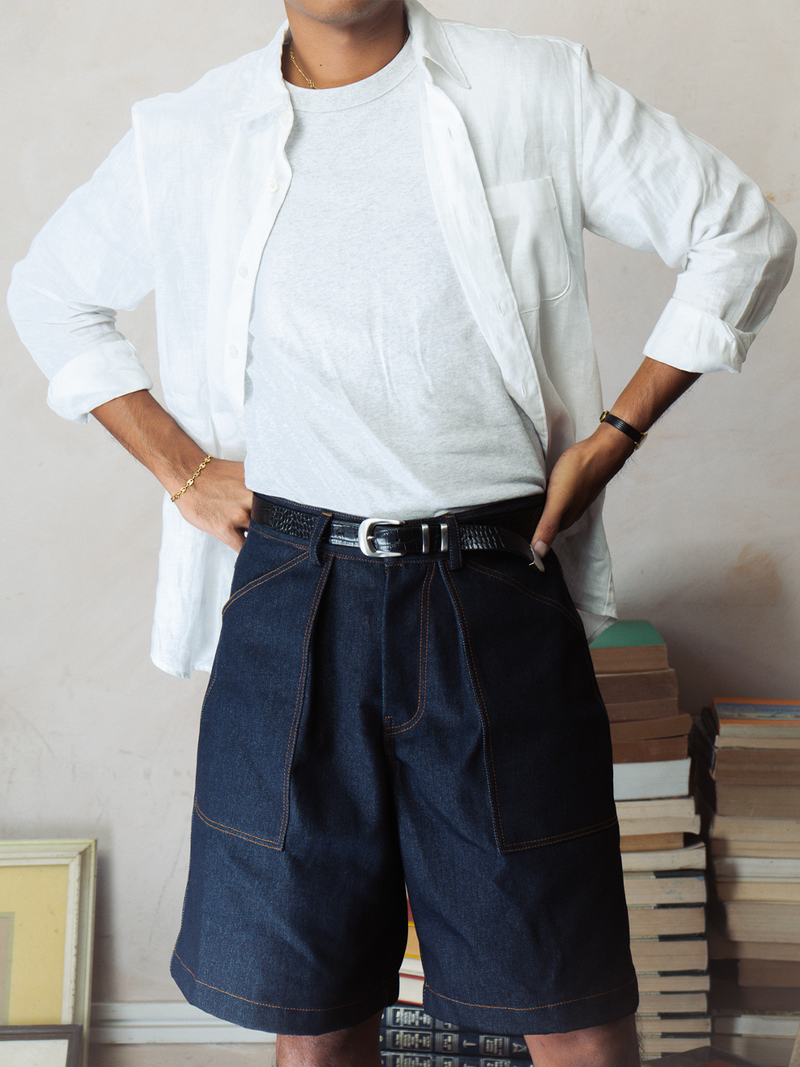Person wearing a white shirt and dark denim shorts with a belt, standing against a neutral wall.