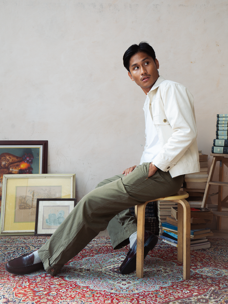 Man sitting on a stool in a room with books and framed pictures wearing a tous cream type 2 trucker jacket. 
