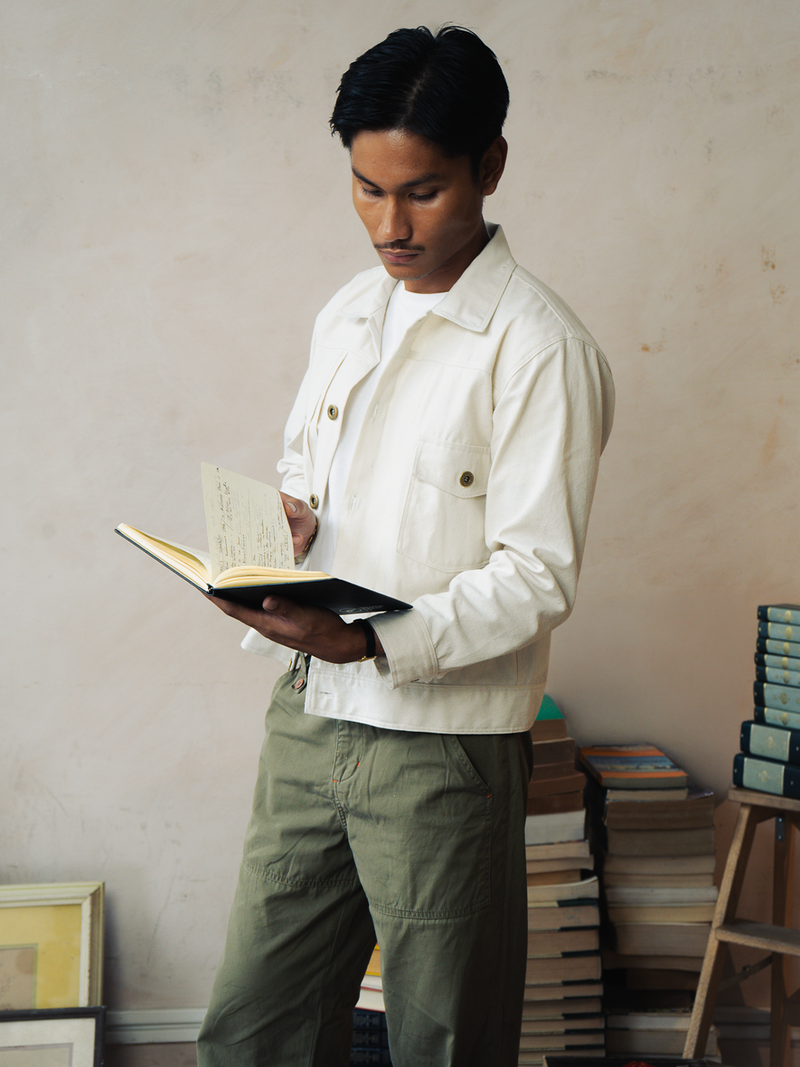 Man holding a book in a room with books stacked on shelves wearing a canvas type 2 jacket. 