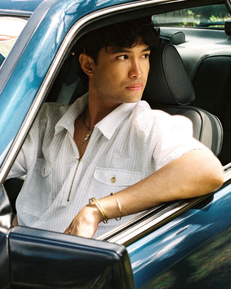 Man sitting in a car wearing a white seer sucker mechanic shirt and gold bracelet.