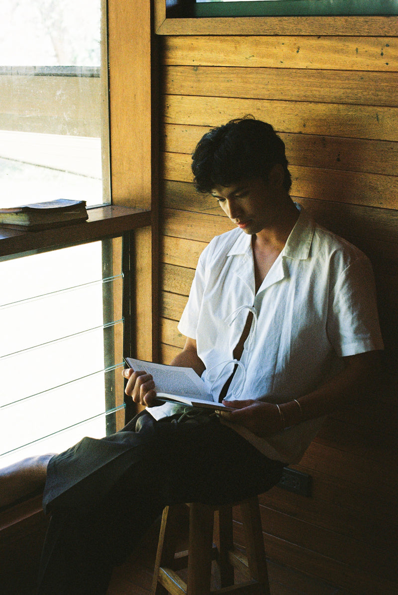 Person sitting on a stool reading a book in a sunlit room with wooden walls.