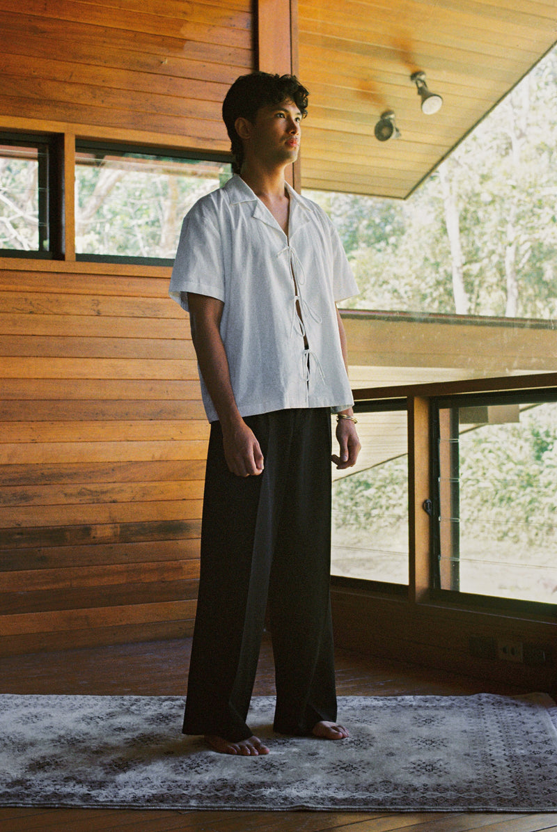 Man standing in a room with wooden walls and large windows, wearing a white lace up sako shirt and dark pants.