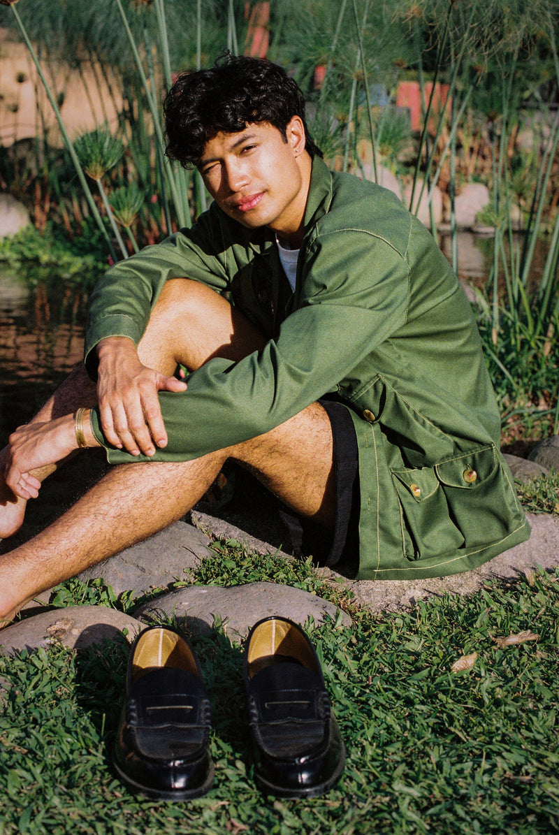 Person wearing a green gardening jacket sitting on grass with shoes in front