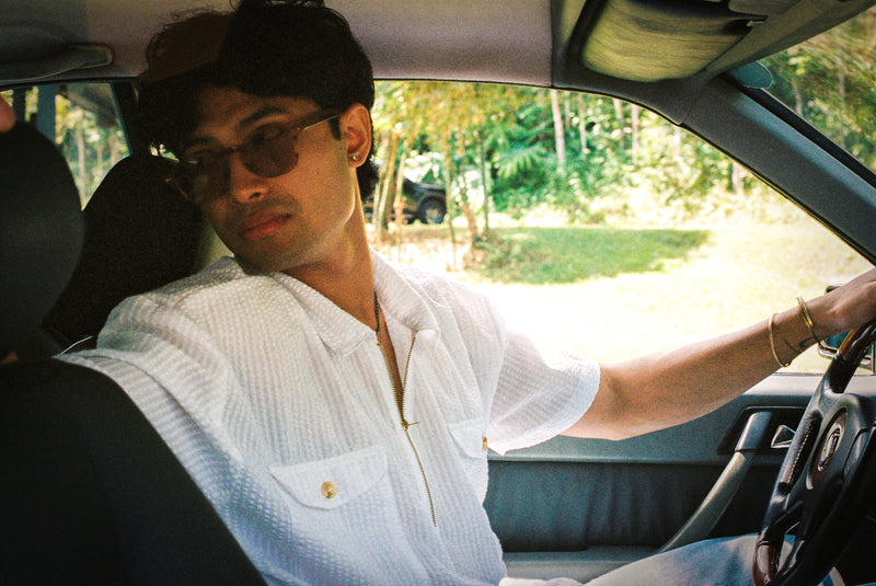 Person sitting inside a car wearing sunglasses and a white seer sucker mechanic shirt.