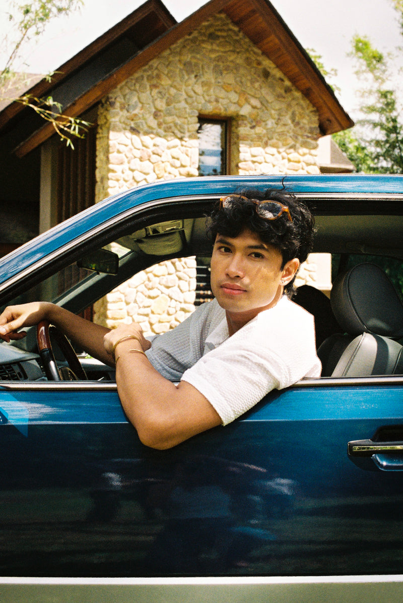 Person sitting in a blue vintage Mercedes Benz car with a stone building in the background wearing a white seer sucker mechanic shirt 