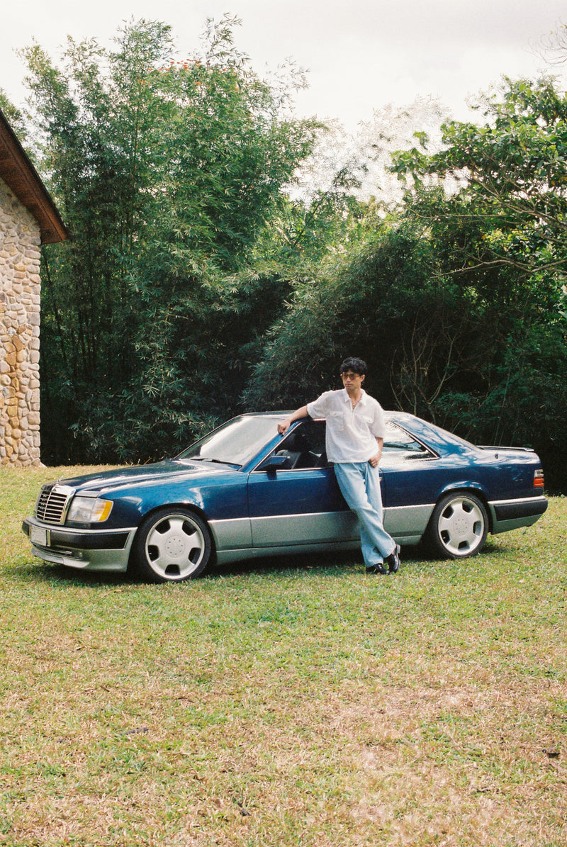 Person standing next to a calssic vintage blue Mercedes-Benz car in a grassy area with trees in the background wearing a white seer sucker mechanic shirt 