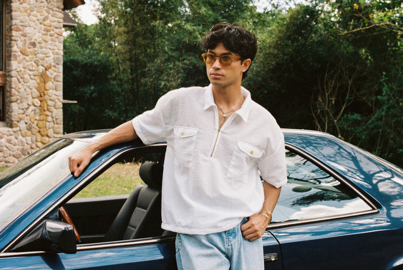 Man in a white seer sucker mechanic shirt and sunglasses leaning against a blue cintage Mercedes Benz outdoors.