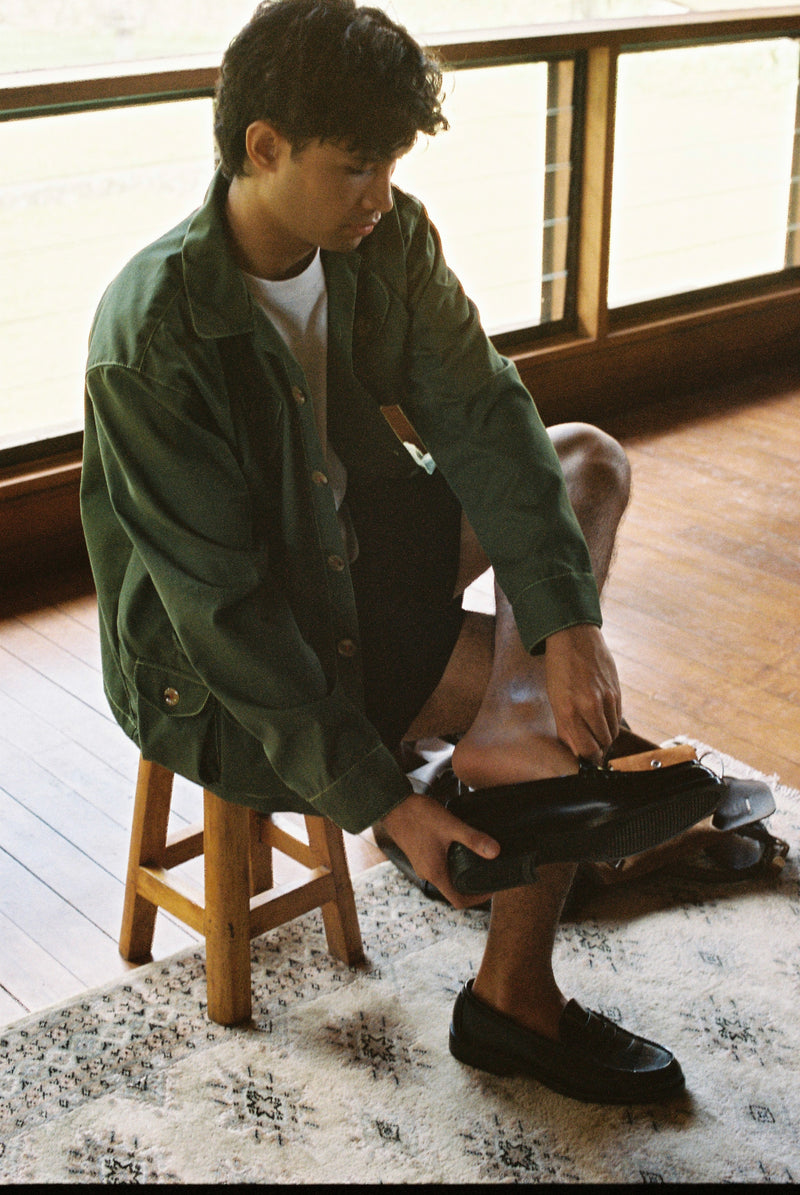 Person sitting on a stool indoors, wearing a green gardening jacket and black shoes.