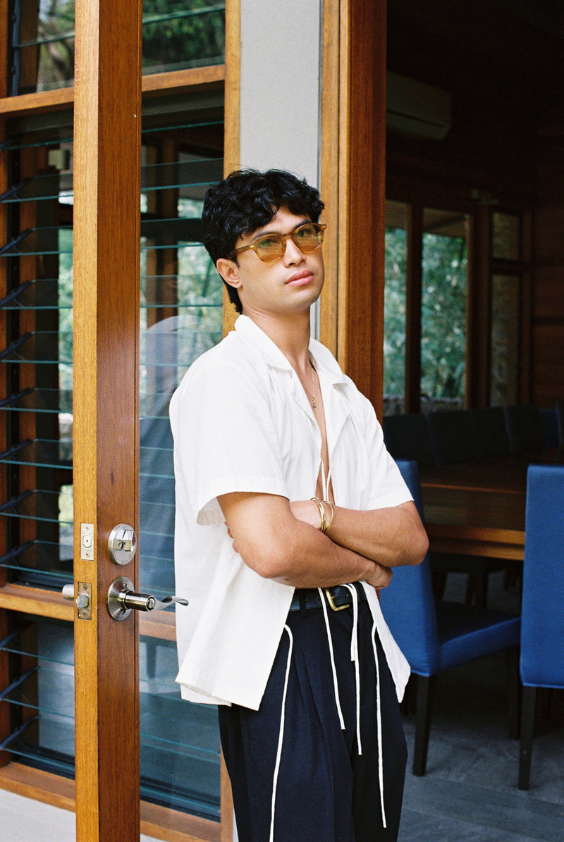Man wearing sunglasses and a lace up sako shirt standing in a room with wooden doors and blue chairs.