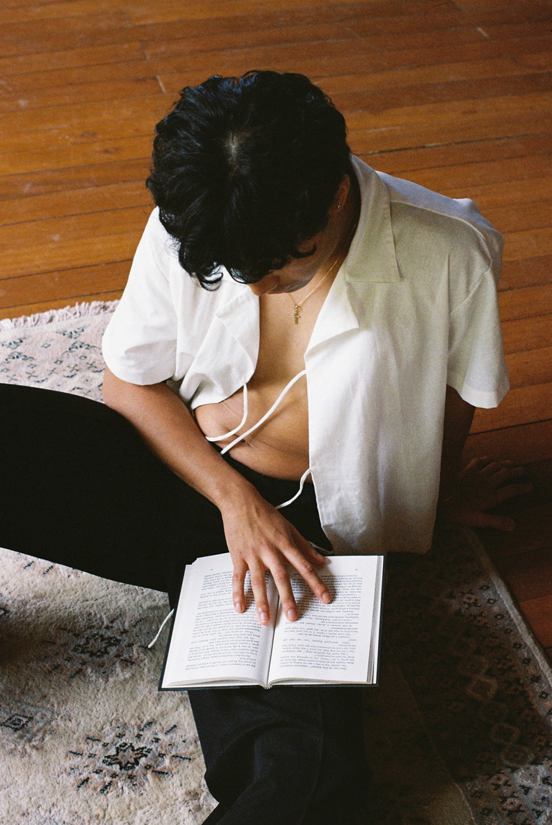 Person sitting on a rug reading a book with a white lace up sako shirt and black pants.