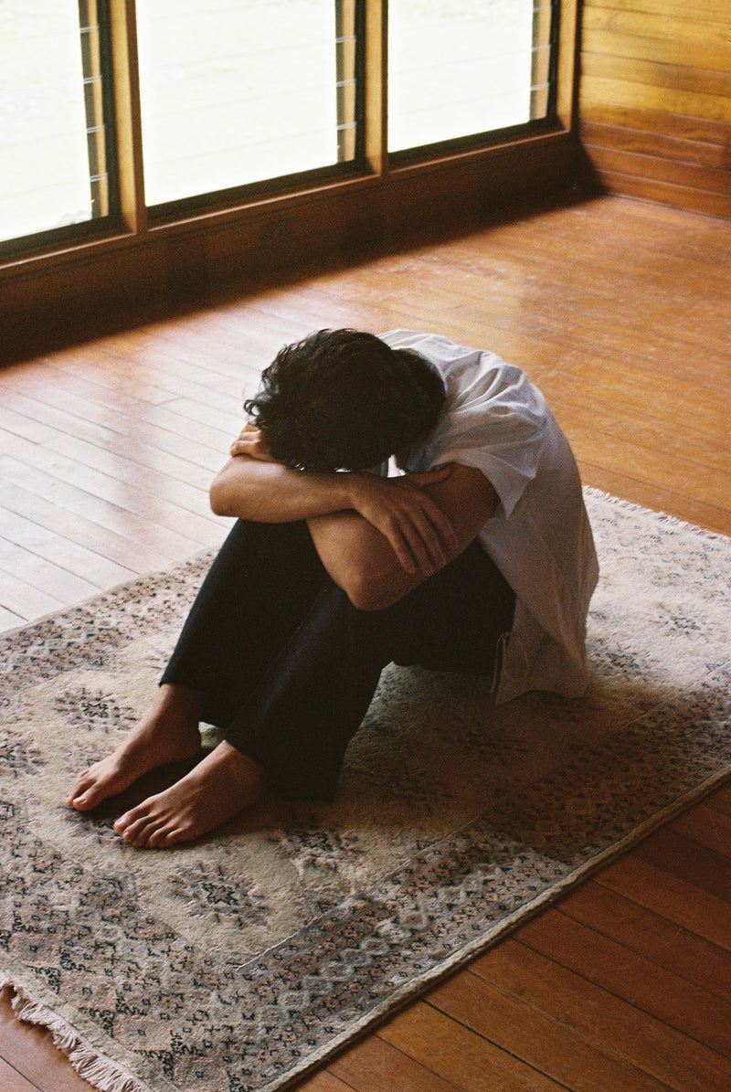 Person sitting on a rug in a room with large windows and wooden floor.