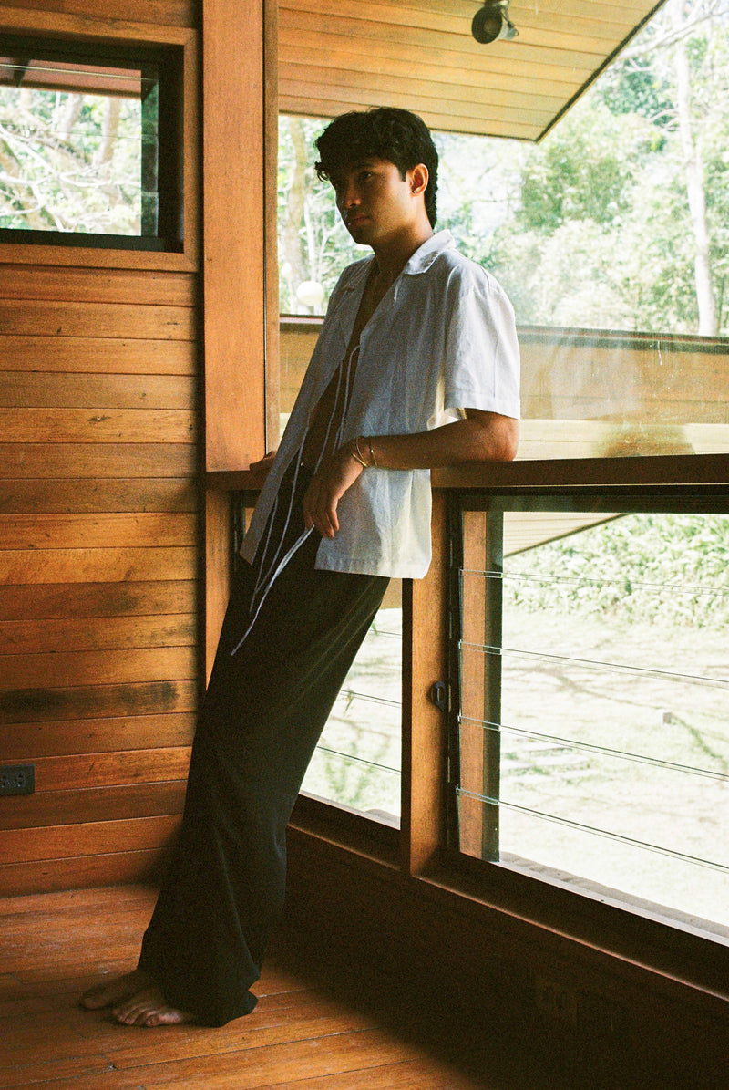Man standing in a modern wooden cabin with large windows wearing a white lace up sako shirt.