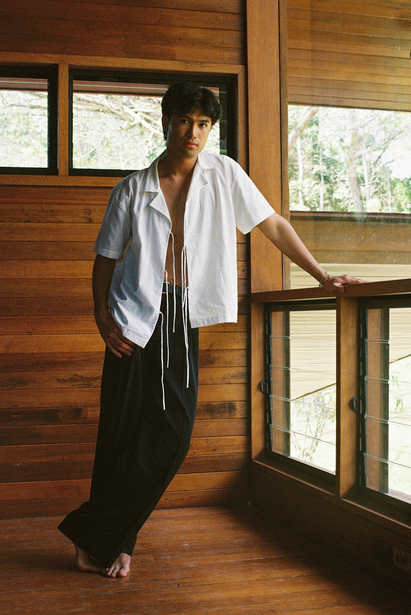 Man wearing a white lace up saki shirt and dark pants standing in a wooden interior.