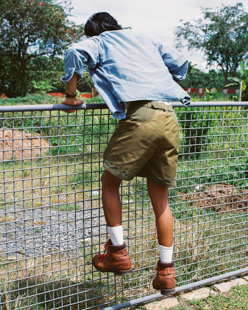 Person wearing a light blue shirt and tous fatigue hiking shorts standing on a wire fence with a garden in the background.