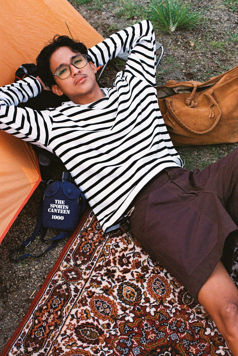 Person lying on a rug with camping gear around, wearing a striped shirt and glasses.