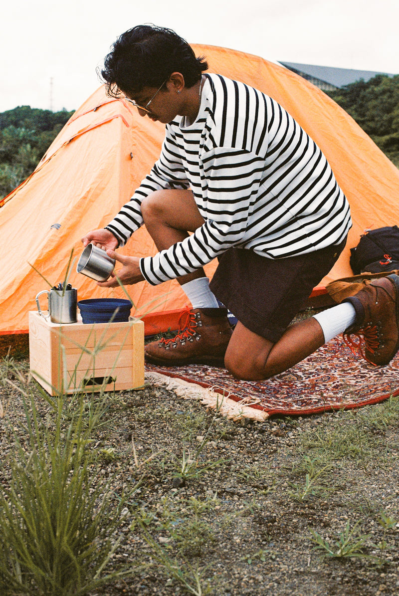 Person camping outdoors, sitting near an orange tent and a wooden crate with a pot.