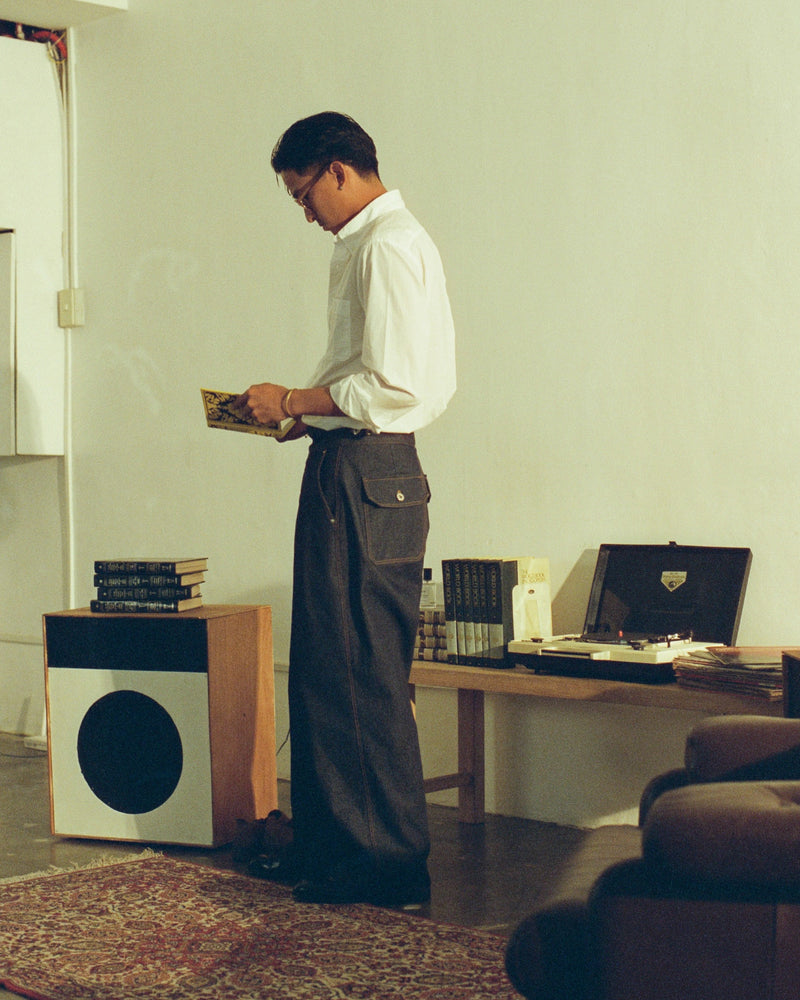 Man in a white shirt and tous denim gurkha pants standing in a room with a desk and computer.