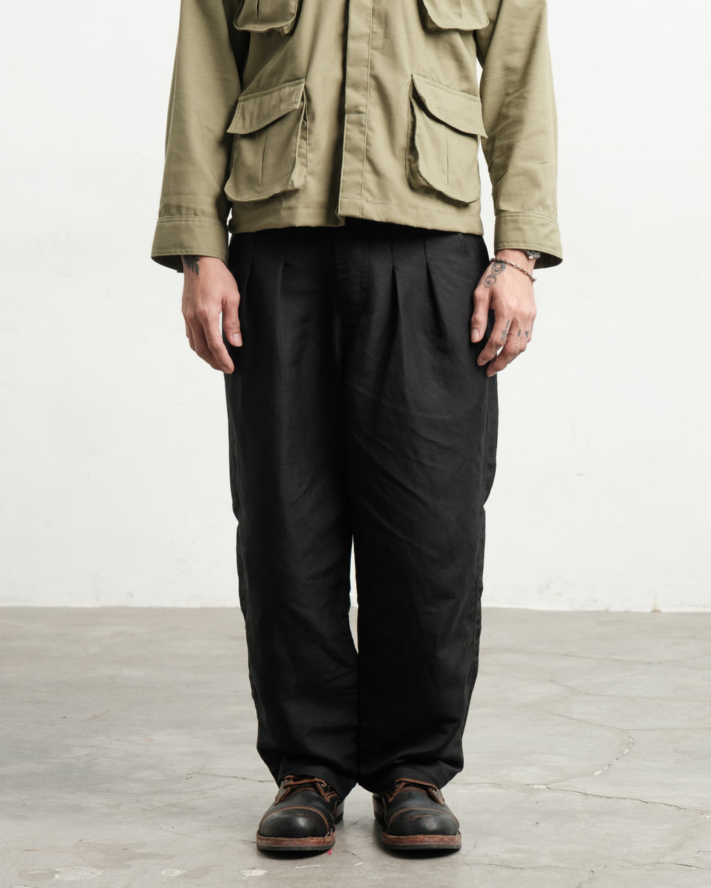 Person wearing a fatigue linen utility workwear jungle jacket and black linen wide legged gurkha pants on a light wooden floor.