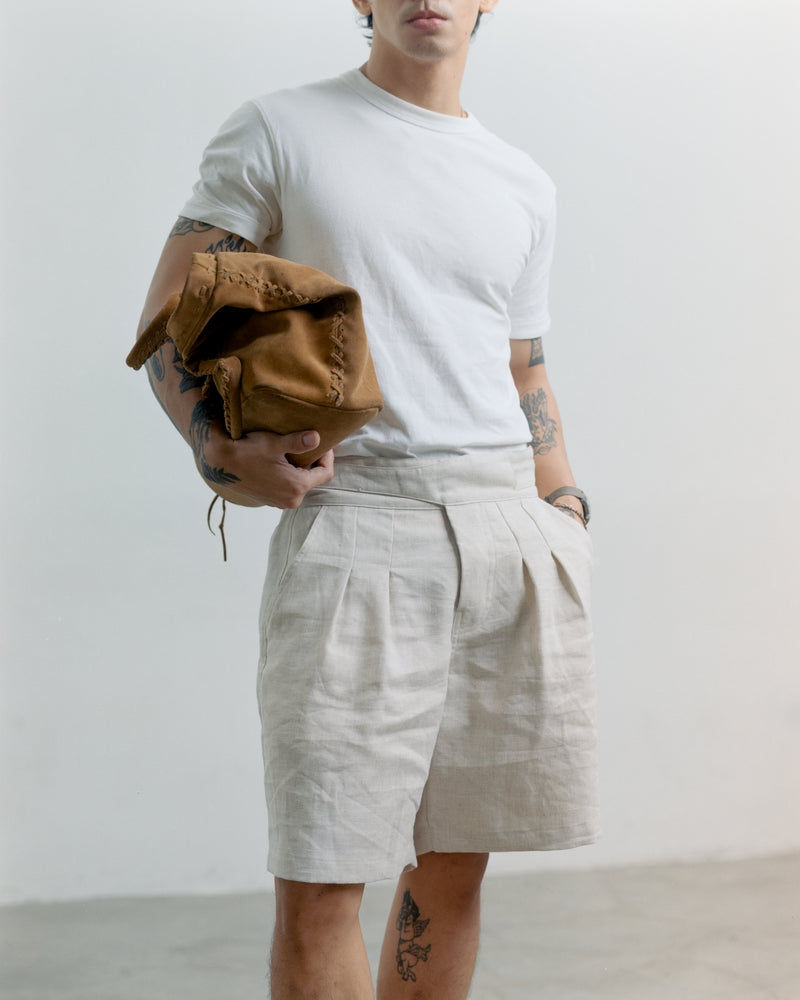 Person wearing a white t-shirt and cream tous linen Gurkha shorts, holding a brown leather bag against a plain background
