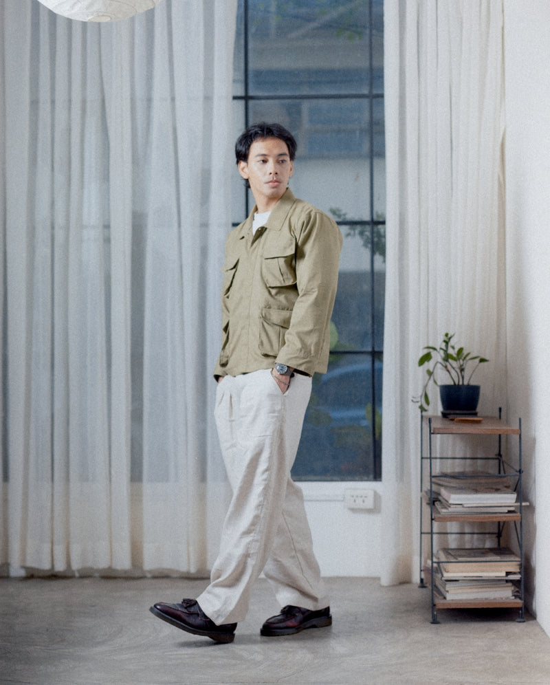 Man standing in a modern room with large windows and minimal decor. Wearing a full linen tous outfit with his fatigue jungle jacket and tous linen cream gurkha pants. 
