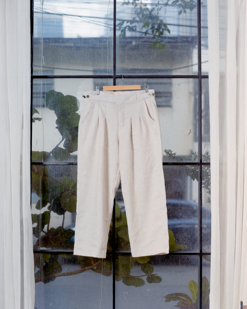 Cream linen gurkha pants hanging on a window in front of a window with a view of trees and buildings.