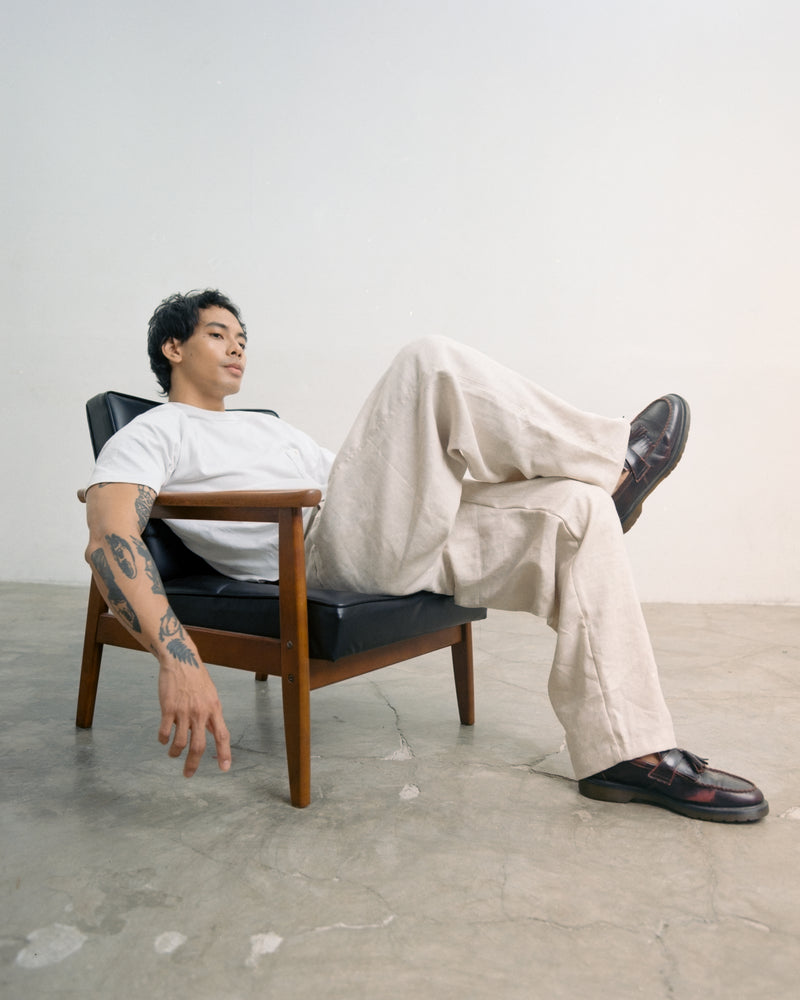 Man sitting on a black chair with a white wall background wearing a tous linen cream gurkha pants. 