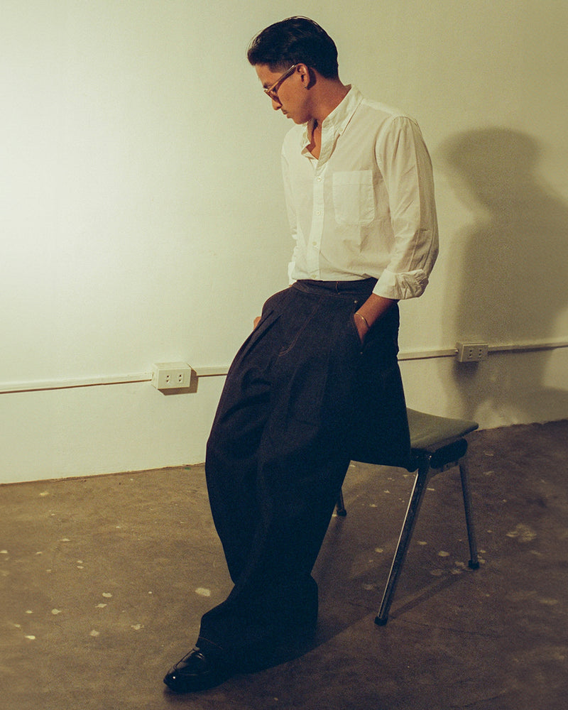 Man in a white shirt and indigo denim gurkha pants standing on a chair against a plain wall.