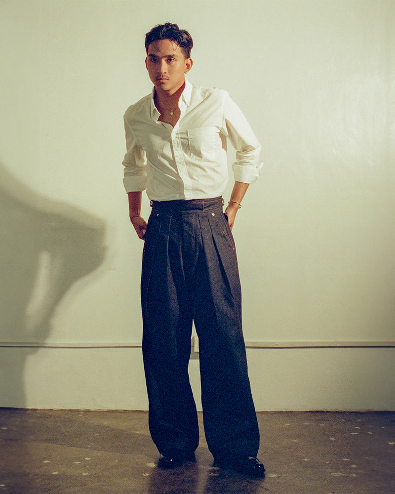 Man wearing a white shirt and denim pleated gurkha pants against a plain background