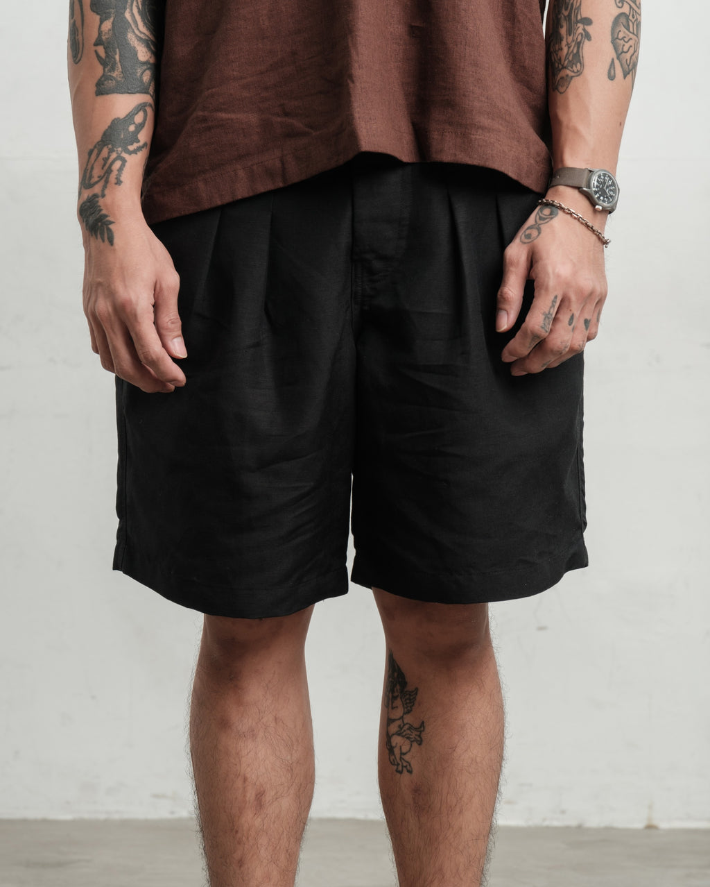 Person wearing black pleated workwear gurkha shorts and a brown shirt against a white wall.