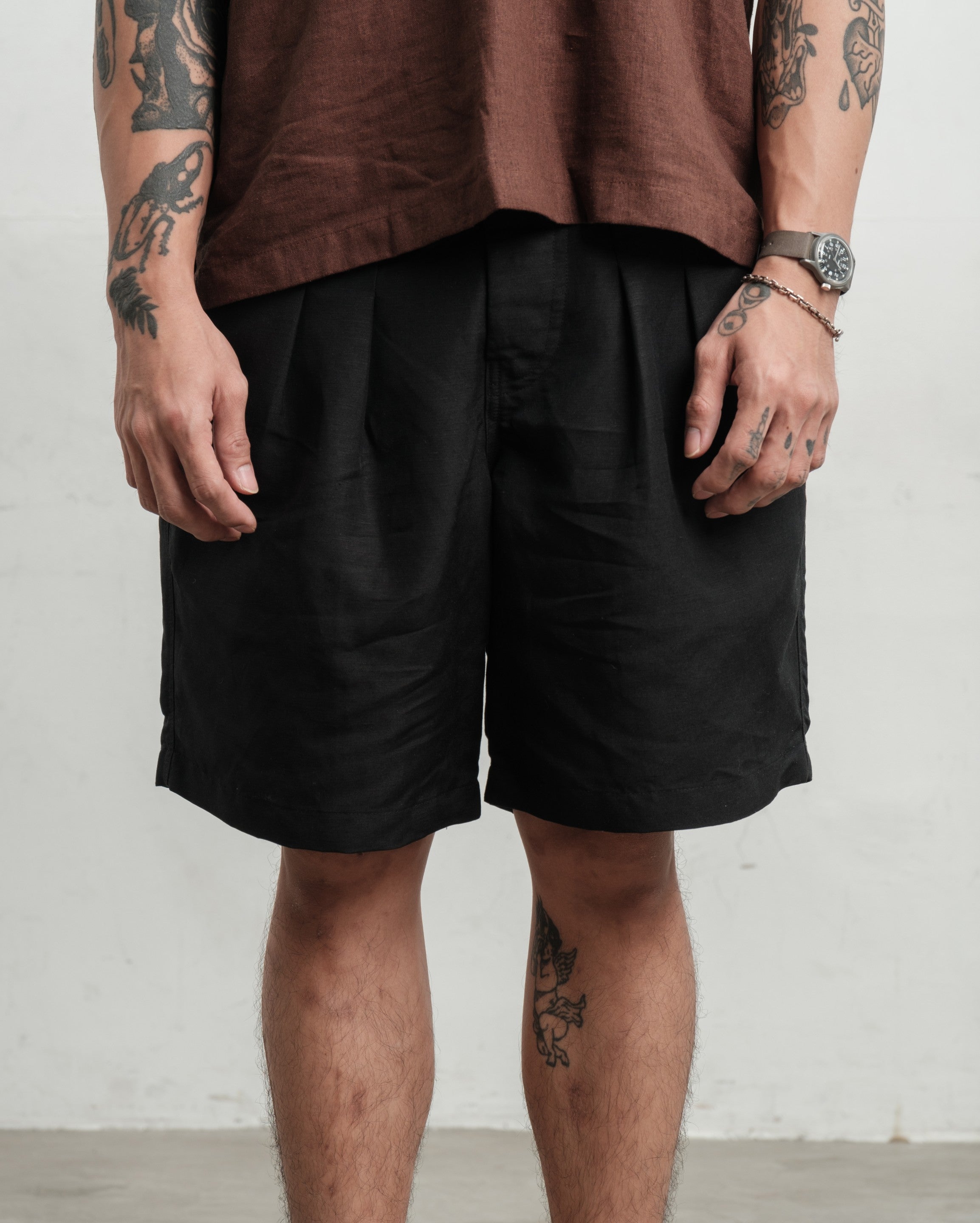 Person wearing black pleated workwear gurkha shorts and a brown shirt against a white wall.