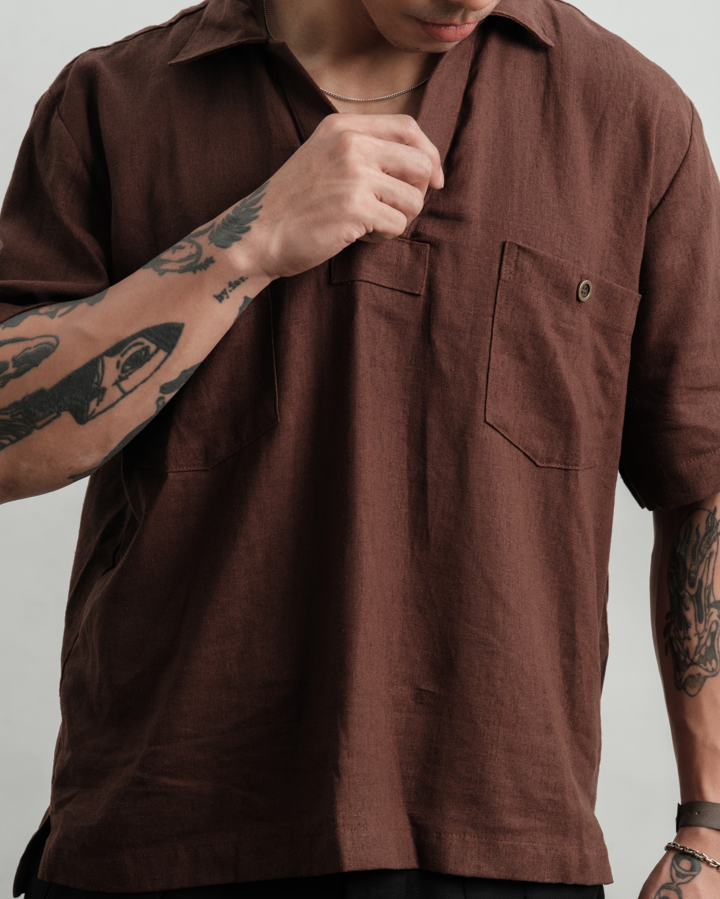 Person wearing arust brown texture linen smock and black linen gurkha shorts with tattoos on arms against a neutral background