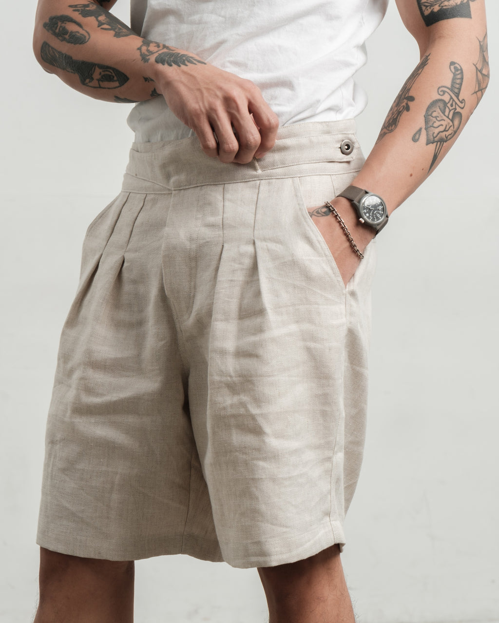 Person wearing cream pleated workwear gurkha shorts with tattoos on arms and a watch on wrist against a plain background