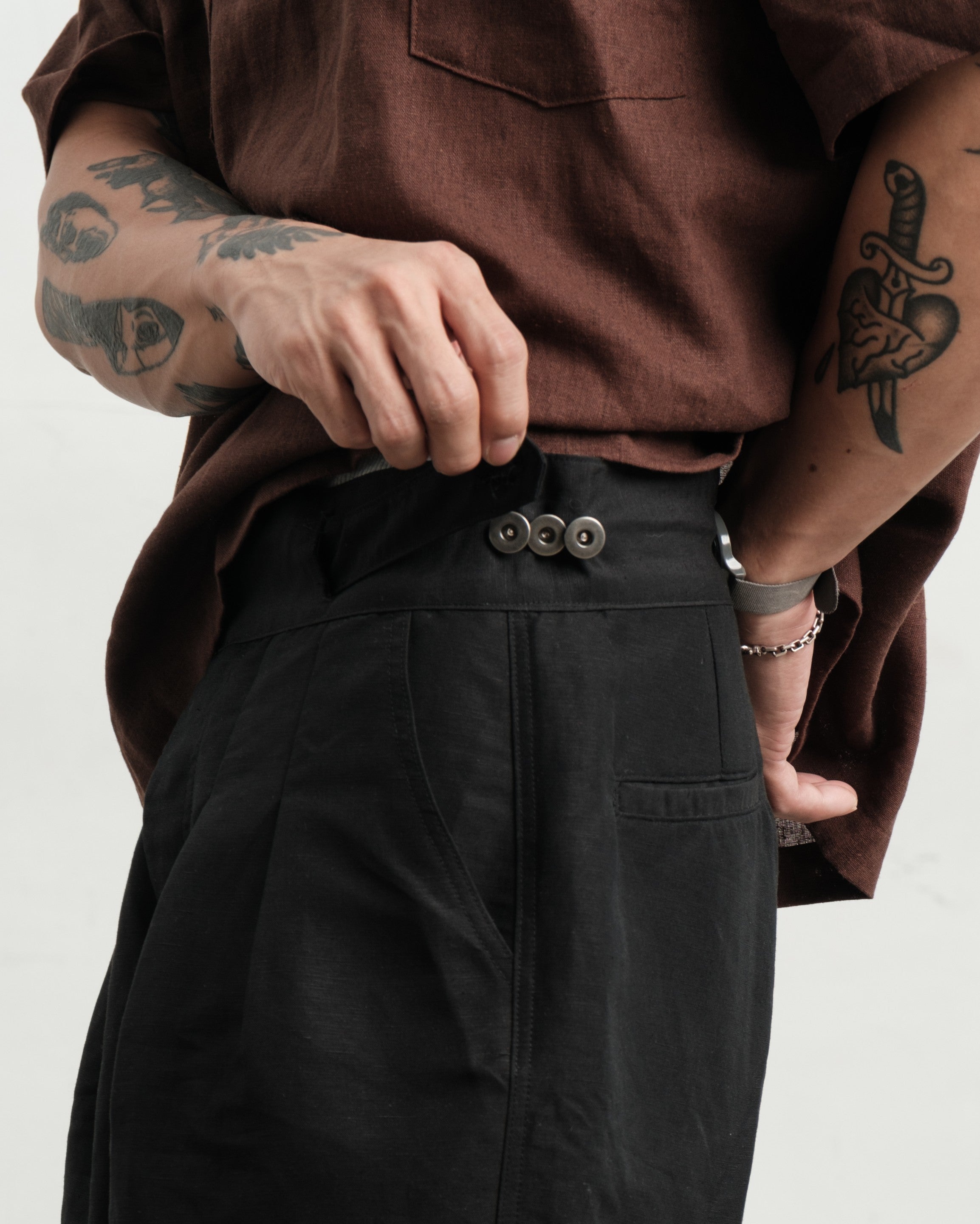 Person wearing a rust brown textured linen smock and black pleated workwear gurkha shorts with visible tattoos on arms, on a white background