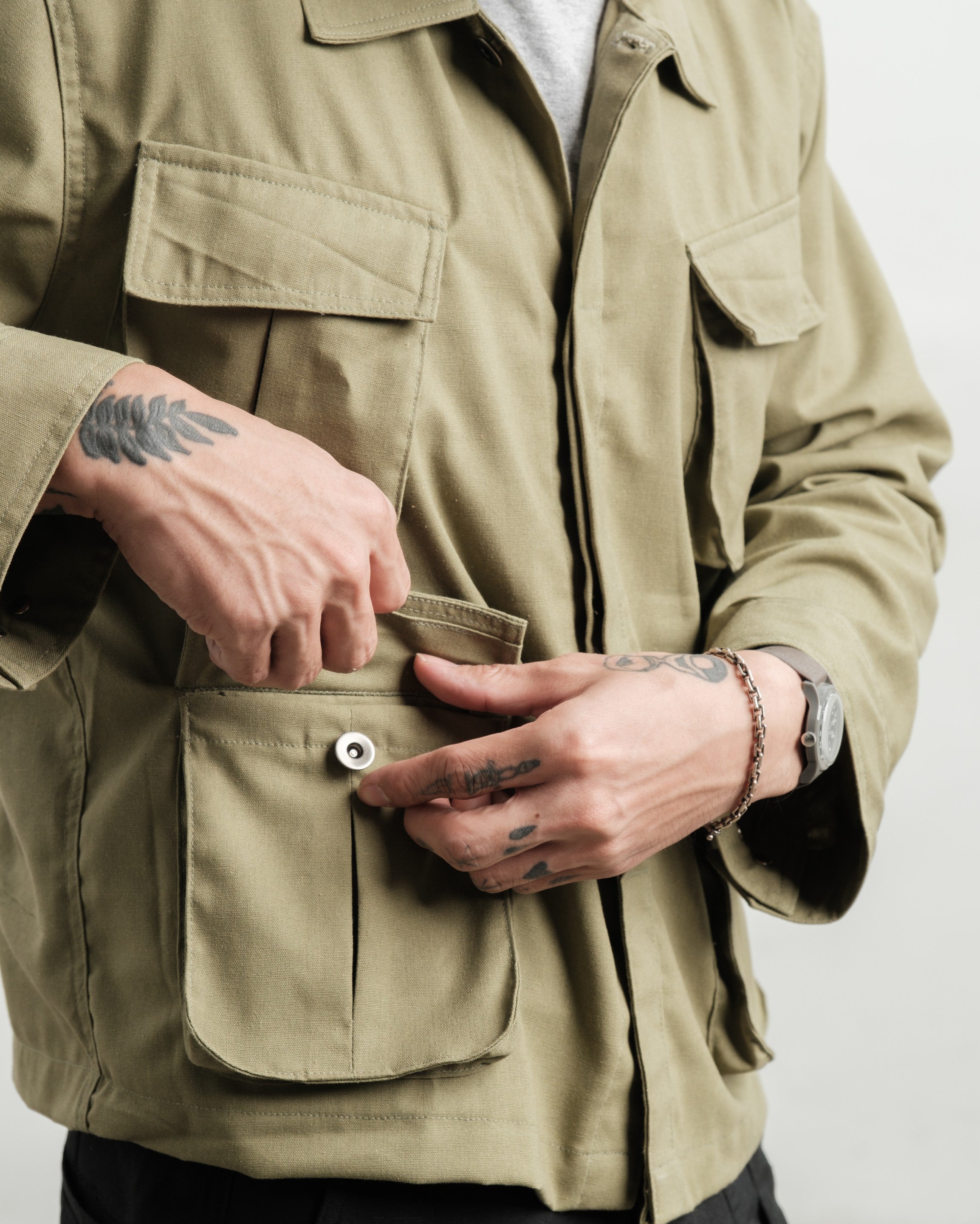 Person wearing a fatigue linen jungle jacket and linen black gurkha pants with visible tattoos on hands and arms opening a pocket.