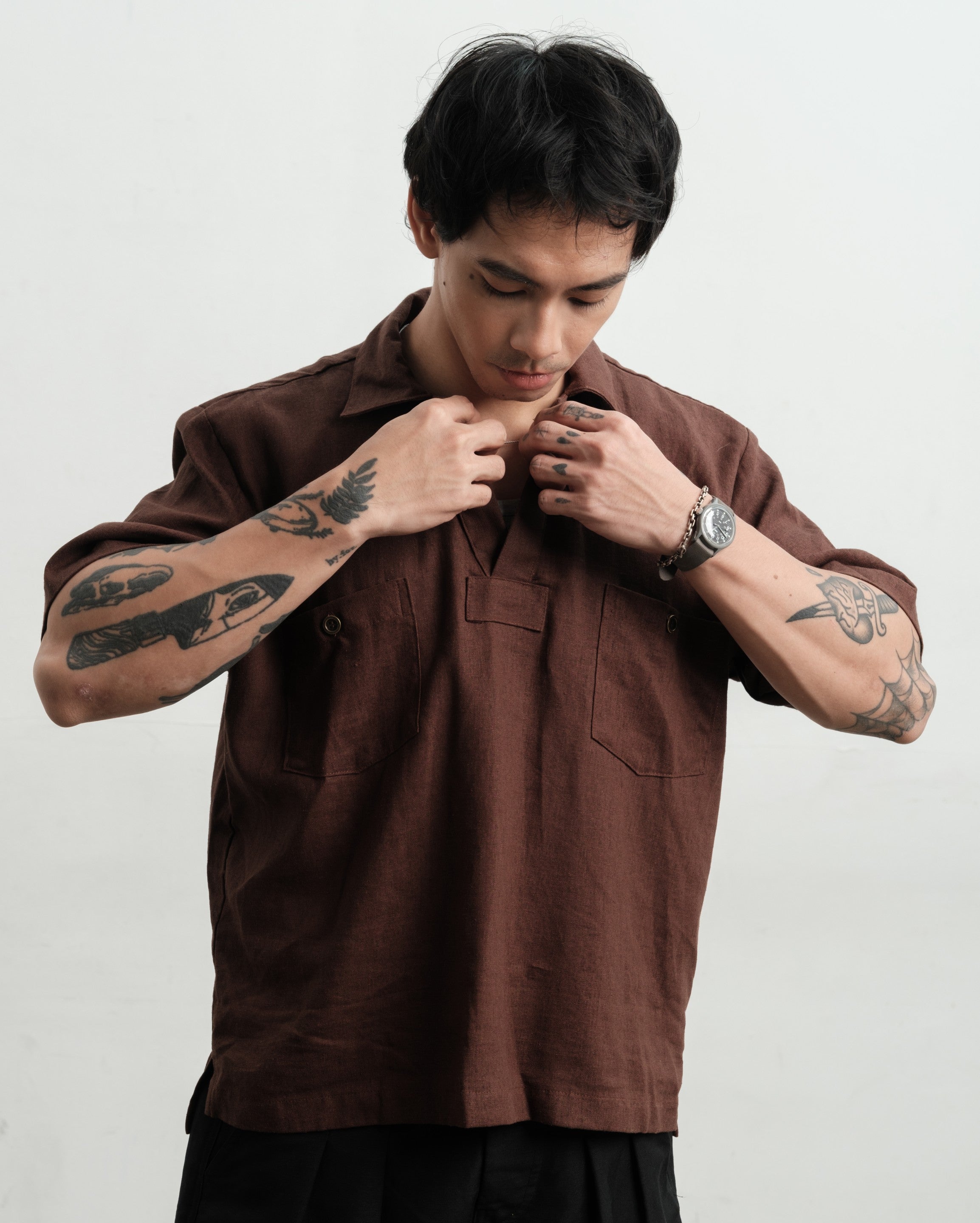 Man wearing a rust brown texture linen smock and black linen gurkha shorts with tattoos on arms against a white background
